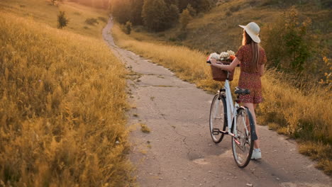 Back-kind-of-Happy-blonde-girl-in-dress-and-hat-turns-around-and-smiling-cheerfully-looks-at-the-camera-and-flirts-strolling-around-the-field-in-summer-with-bike-and-flowers.